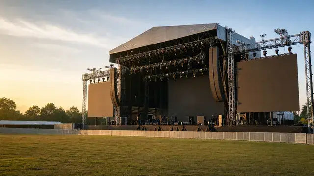 Outdoor festival main stage at golden hour with line array speakers, LED walls, lighting truss, and an empty grass field in front