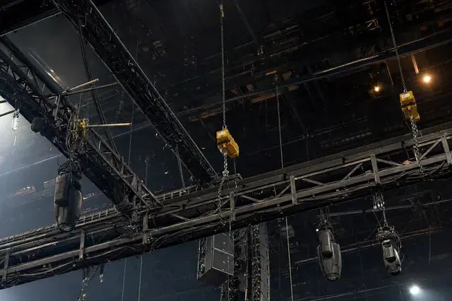 Looking straight up at an arena rigging grid, with steel truss, yellow chain motors, hanging line array, and moving lights silhouetted against work lights
