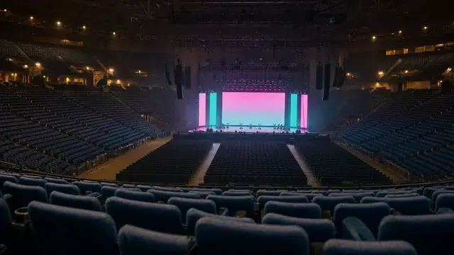 Empty arena an hour before doors, seen from mid-tier seating, with a distant lit stage showing solid magenta and teal LED walls flanked by hanging line arrays