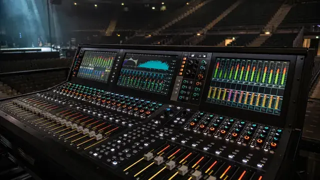 Large digital audio mixing console at a front-of-house position in an empty arena during soundcheck, three touchscreens showing waveform meters and level bars, illuminated faders and encoders across the surface