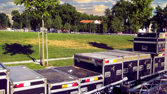 Black road and flight cases with colored labels stacked on a grassy park lawn under a summer evening sky, waiting between shows
